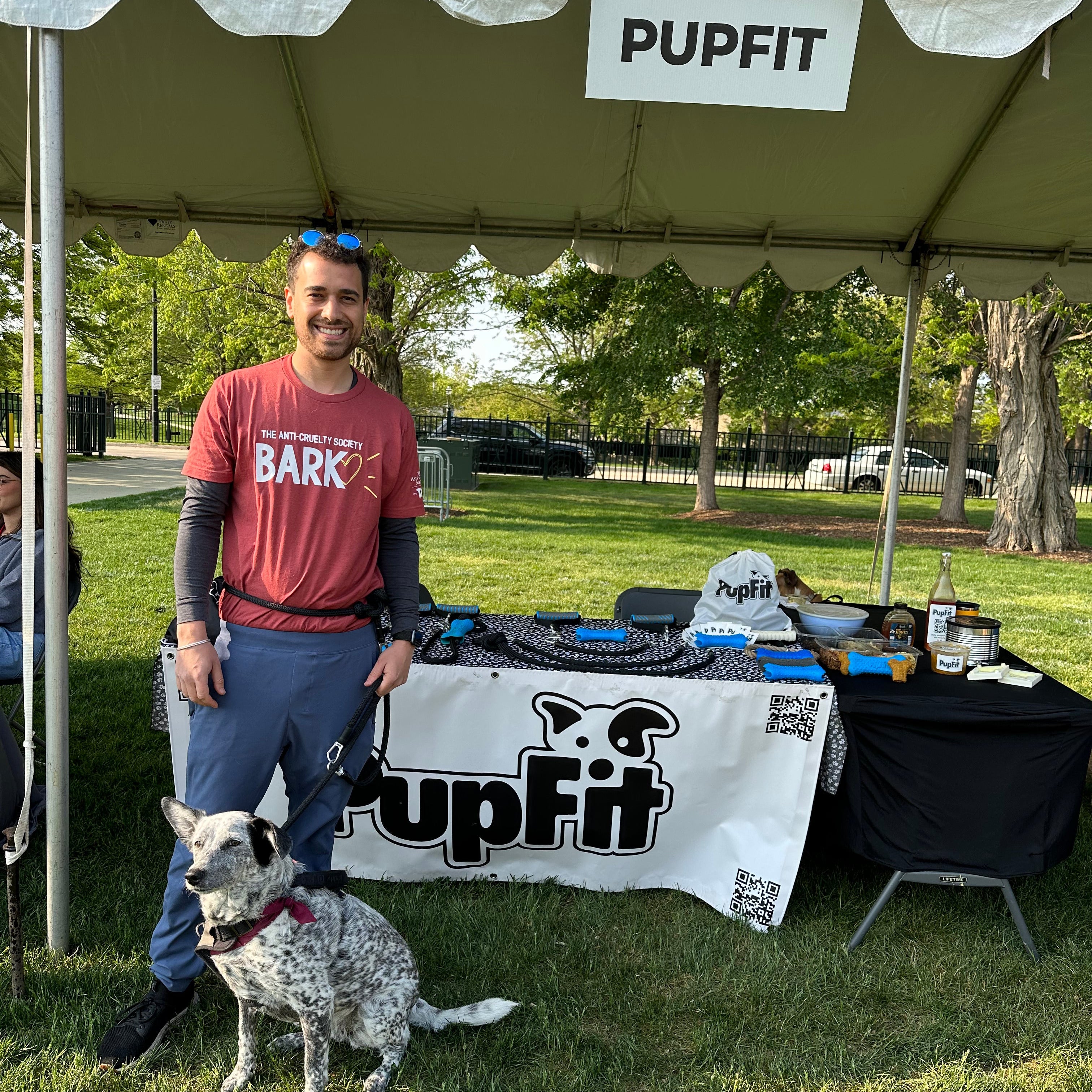 Man with a dog standing under a 'PUPFIT' tent in a park setting.