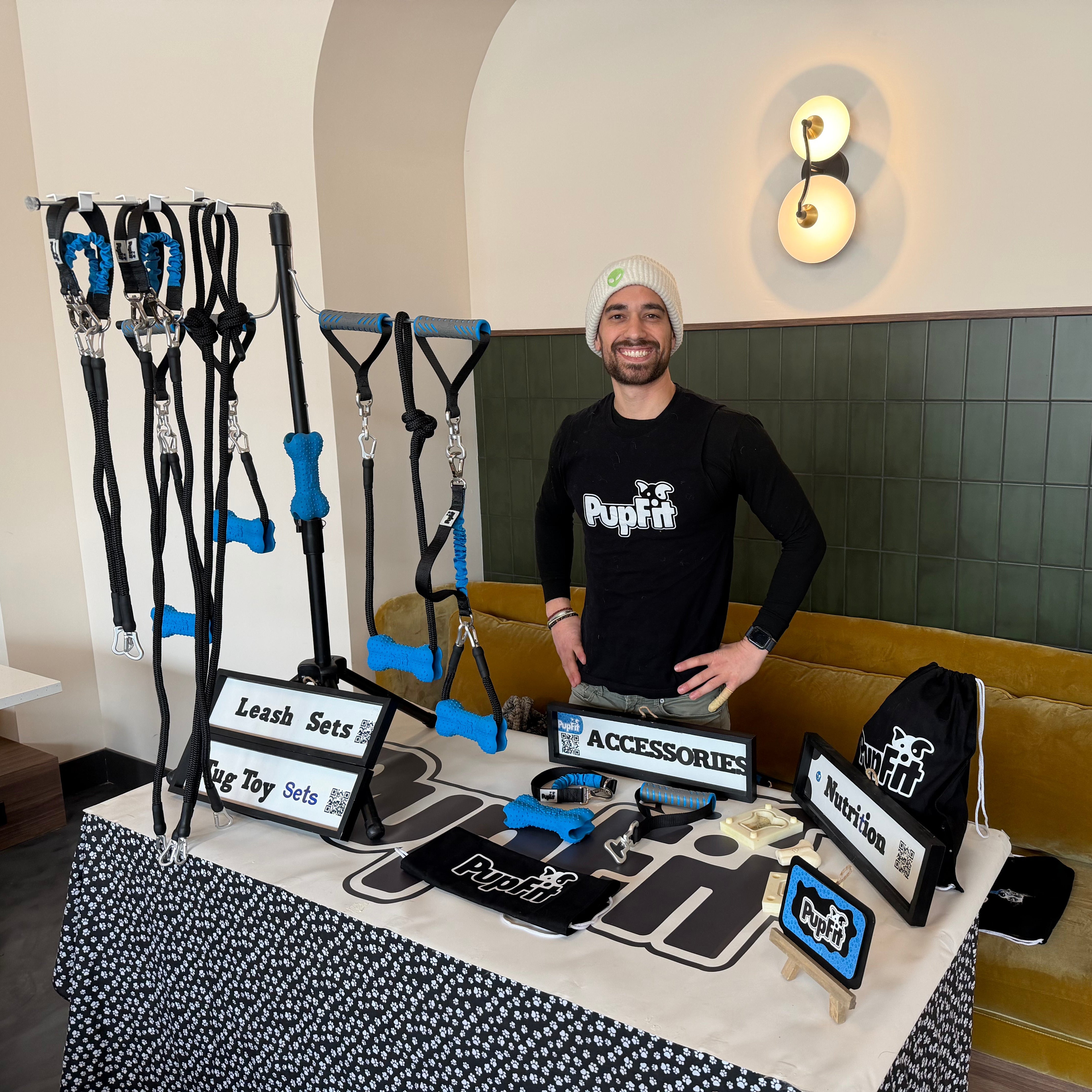 Man standing behind a PupFit table displaying PupFit products with a couch and wall decor in the background.
