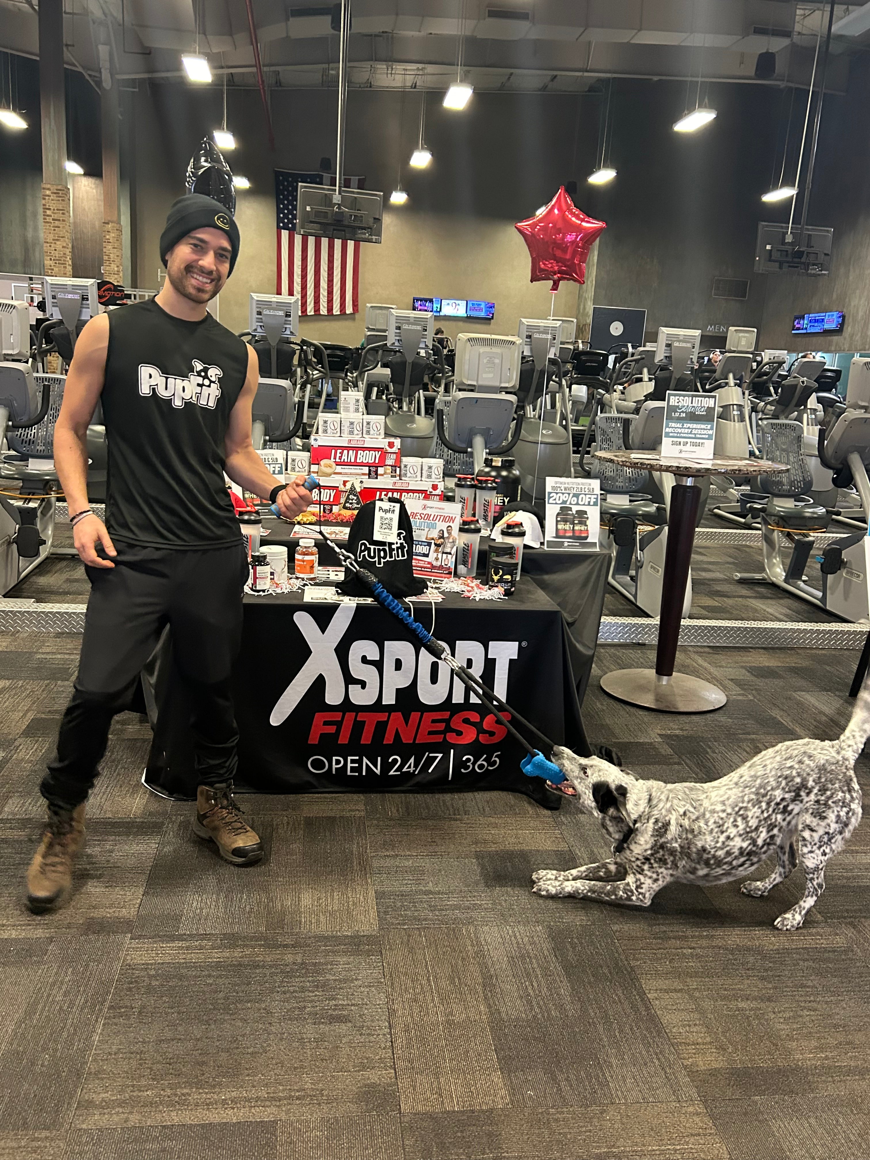 Man in a gym with a dog, standing next to an XSport Fitness table with PupFit gear.